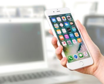 Hand holding a smartphone displaying app icons, with a laptop in the background
